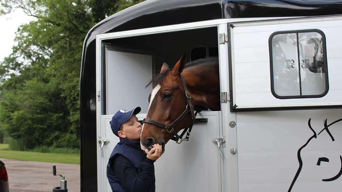 A horse sticks its head out of a van. A young boy kisses him on the nose.