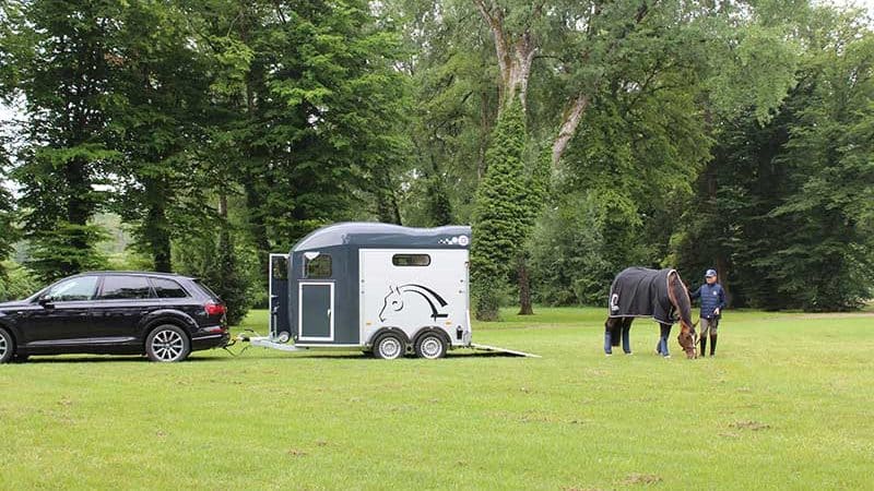 A van hitched to a car in a green meadow. A horse and rider graze nearby.