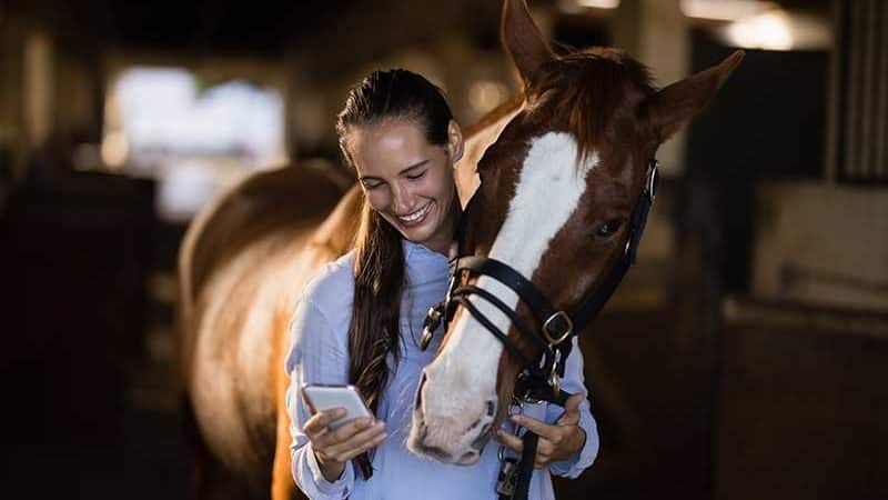 Young woman looking at her smartphone and smiling. His horse looks over his shoulder.